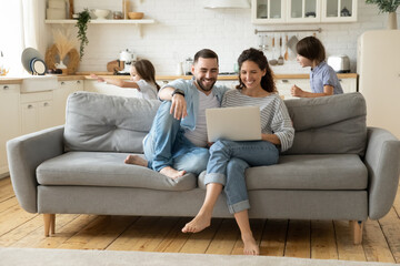 Happy parents using laptop, sitting on couch while kids playing funny game, smiling mother and father shopping online, enjoying leisure time, little son and daughter running and laughing together
