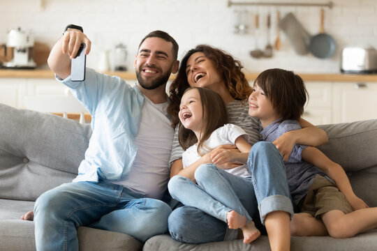 Happy Parents With Adorable Little Kids Having Fun, Using Mobile Device, Smiling Father Holding Smartphone, Taking Selfie, Overjoyed Young Family With Daughter And Son Sitting On Cozy Couch At Home