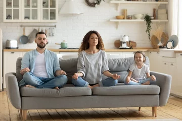 Poster Im Rahmen Peaceful young mother and father with little daughter practicing yoga together, sitting on cozy couch in lotus pose, calm satisfied parents with kid meditating, relaxing, enjoying leisure time © fizkes