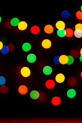 Multicolored bokeh from garlands on a black background. A versatile festive and magical backdrop. Christmas, new year, halloween. Red, yellow, green, blue circles glow in dark. Vertical, out of focus