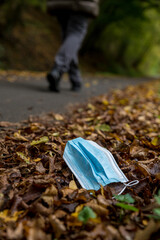 A discarded protective face mask lying next to a road on brown, autumnal leaves. A person is walking by.