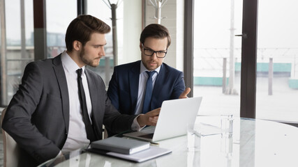 Focused young male business partners in suits sitting at table, looking at computer screen,...