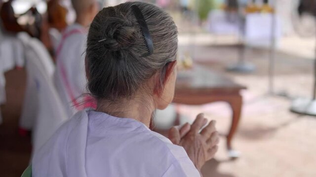 An old Thai Buddhist woman dressed in a white cloth is raising her hands while attending a religious practice at a Buddhist temple.