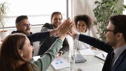 Positive happy young multiracial diverse colleagues giving high five, celebrating group business achievement, company growth or financial marketing strategy results at brainstorming meeting in office.