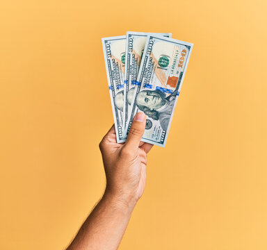 Hand of hispanic man holding 100 dollars banknotes over isolated yellow background.
