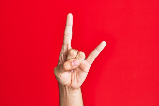 Arm of caucasian white young man over red isolated background gesturing rock and roll symbol, showing obscene horns gesture