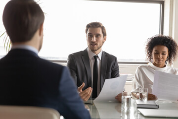 Back rear view confident team leader listening to young male colleague reporting sales or explaining marketing research results at briefing meeting with multiracial coworkers in modern office.
