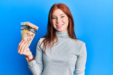 Young red head girl holding canadian dollars looking positive and happy standing and smiling with a confident smile showing teeth