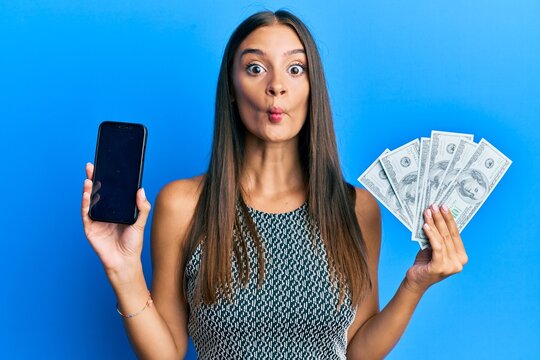 Young hispanic woman holding dollars and using smarpthone making fish face with mouth and squinting eyes, crazy and comical.