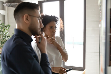 Side view focused young african american employee thinking of difficult problem solution with arabian ethnicity colleague, managing workflow or planning strategy on glass wall in modern office.