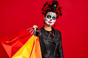 Girl with sugar skull makeup with a wreath of flowers on her head and skull, wearth lace gloves and leather jacket, hold shopper bags party preparation isolated on red background