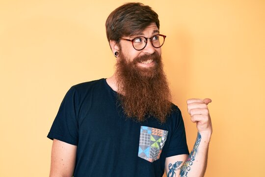Handsome young red head man with long beard wearing casual clothes and glasses smiling with happy face looking and pointing to the side with thumb up.
