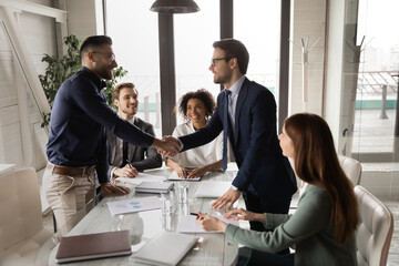 Happy young multiracial diverse business people in formal wear watching two male confident partners shaking hands, establishing profitable cooperation, celebrating successful collaboration at meeting. - Powered by Adobe