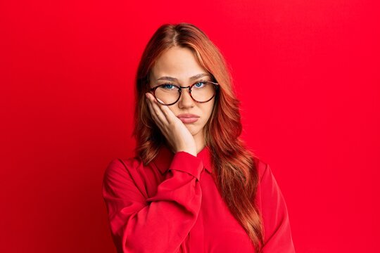 Young beautiful redhead woman wearing casual clothes and glasses over red background thinking looking tired and bored with depression problems with crossed arms.