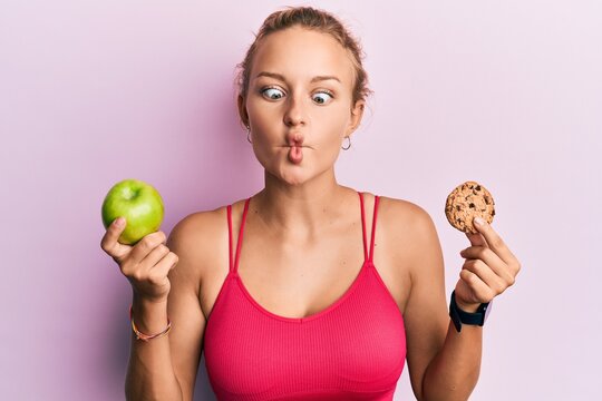 Beautiful caucasian woman holding green apple and cookie making fish face with mouth and squinting eyes, crazy and comical.