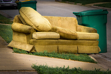 Old yellow couch with cushions sits on a curb in residential area next to a recycling cart waiting...