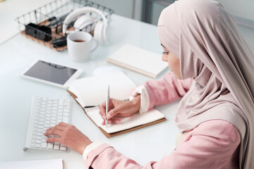 Contemporary young Muslim woman in hijab and smart casualwear making notes