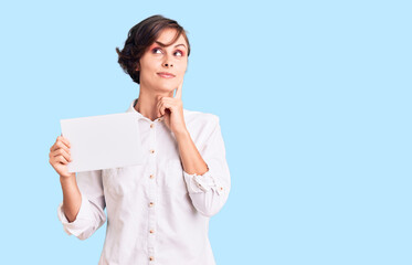 Beautiful young woman with short hair holding blank empty banner serious face thinking about question with hand on chin, thoughtful about confusing idea