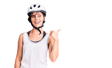 Beautiful young woman with short hair wearing bike helmet smiling with happy face looking and pointing to the side with thumb up.