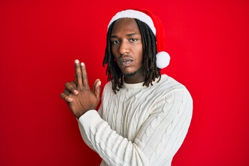 African american man with braids wearing christmas hat holding symbolic gun with hand gesture, playing killing shooting weapons, angry face