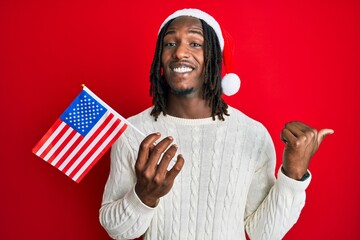African american man with braids wearing christmas hat and usa flag pointing thumb up to the side smiling happy with open mouth