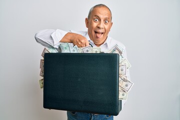 Handsome senior man holding briefcase full of dollars sticking tongue out happy with funny expression.