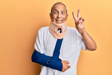 Handsome mature senior man wearing cervical collar and arm on sling smiling with happy face winking at the camera doing victory sign with fingers. number two.