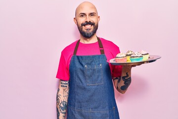 Young handsome man wearing apron holding cupcake looking positive and happy standing and smiling with a confident smile showing teeth