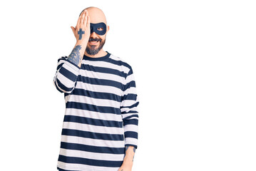 Naklejka premium Young handsome man wearing burglar mask covering one eye with hand, confident smile on face and surprise emotion.