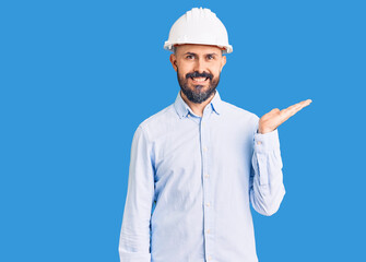 Young handsome man wearing architect hardhat smiling cheerful presenting and pointing with palm of hand looking at the camera.