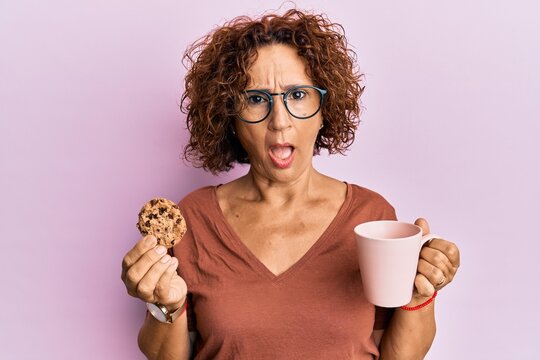 Beautiful Middle Age Mature Woman Drinking A Cup Of Coffee And Cookie In Shock Face, Looking Skeptical And Sarcastic, Surprised With Open Mouth