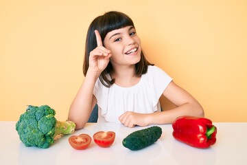 Young little girl with bang sitting on the table with veggies smiling with an idea or question pointing finger up with happy face, number one