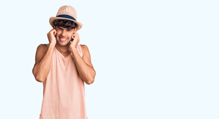 Young hispanic man wearing summer hat covering ears with fingers with annoyed expression for the noise of loud music. deaf concept.