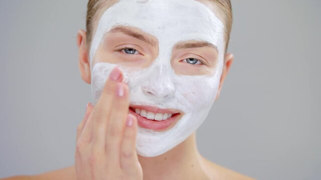 Close-Up Beauty Portrait Of Young Caucasian Blonde Woman With Blue Eyes She Applying White Mask On Face And Smiling On Gray Background She Using Hand And Finger On Healthy Skin Care Concept Slow Mo