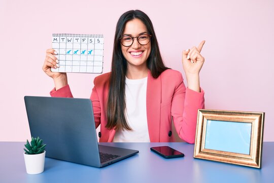 Young caucasian woman sitting at the desk holding travel calendar smiling happy pointing with hand and finger to the side