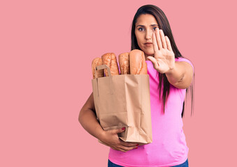 Young beautiful brunette woman holding delivery bag with bread with open hand doing stop sign with serious and confident expression, defense gesture