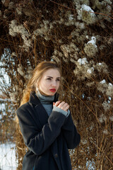 Fashion portrait of pretty woman with red lipstick wearing a dark coat against winter nature background.