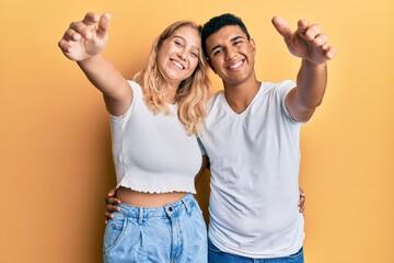 Young interracial couple wearing casual white tshirt looking at the camera smiling with open arms for hug. cheerful expression embracing happiness.