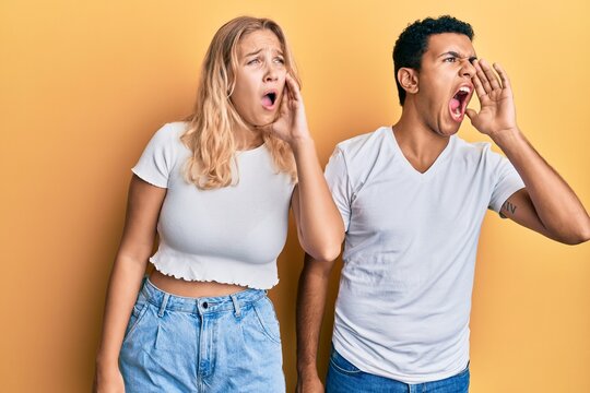 Young interracial couple wearing casual white tshirt shouting and screaming loud to side with hand on mouth. communication concept.