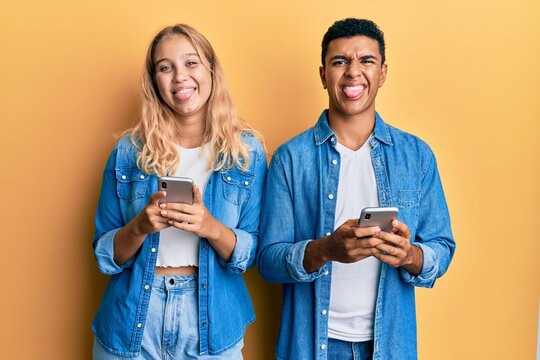 Young interracial couple using smartphone sticking tongue out happy with funny expression.