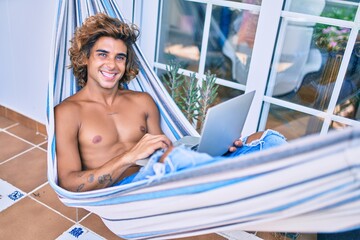 Young hispanic man relaxed working using laptop lying on the hammock at terrace.