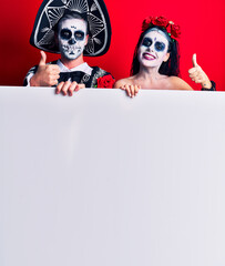 Young couple wearing mexican day of the dead costume holding blank empty banner smiling happy and positive, thumb up doing excellent and approval sign