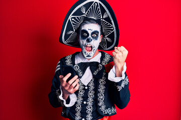 Young man wearing mexican day of the dead costume using smartphone screaming proud, celebrating victory and success very excited with raised arms