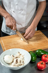 Hands of young male chef cutting fresh onions and champignons on wooden board