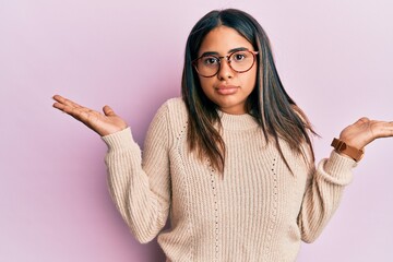 Young latin girl wearing casual winter sweater and glasses clueless and confused with open arms, no...