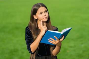 Serious little kid with thoughtful look hold school book green grass sunny summer outdoors, imagination