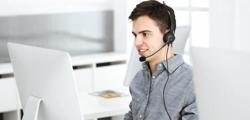 Naklejka premium Casual dressed young man using headset and computer while talking with customers online. Call center, business concept