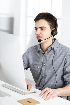 Casual dressed young man using headset and computer while talking with customers online. Call center, business concept