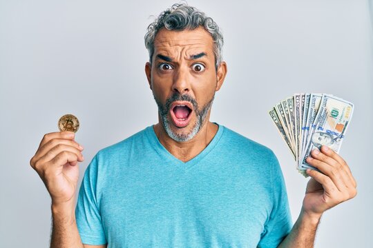 Middle age grey-haired man holding dollars and bitcoin afraid and shocked with surprise and amazed expression, fear and excited face.