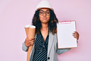 Young african american woman wearing architect hardhat holding blueprints and clipboard puffing cheeks with funny face. mouth inflated with air, catching air.
